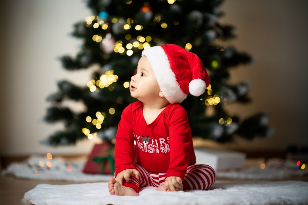 A baby wearing a Santa suit sits happily in front of a decorated Christmas tree.  This image is for a blog post about baby's first Christmas, with gift ideas and traditions for new parents.